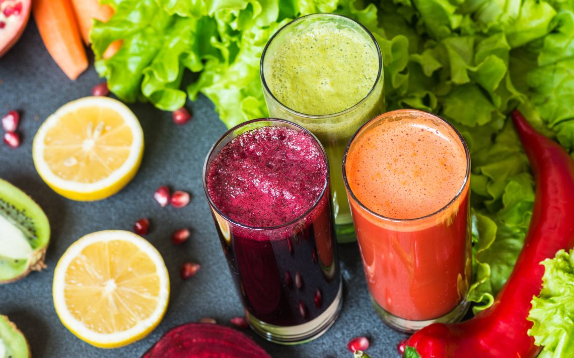 Three glasses of fresh juice surrounded by fruits and vegetables